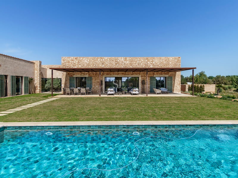Exterior de villa de lujo con piscina turquesa, césped y fachada de piedra natural bajo un cielo despejado.