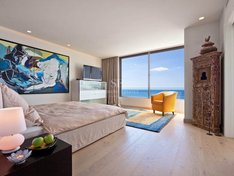 Chambre moderne avec vue sur mer, grand lit, fauteuil jaune et armoire en bois sculpté.