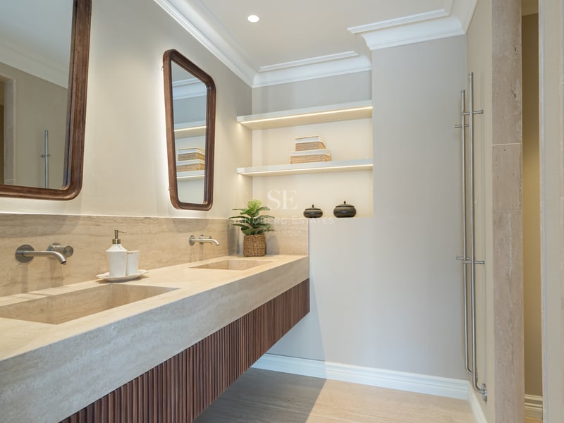 Modern bathroom featuring a dual travertine stone vanity, wood-slat cabinets, and LED-lit recessed shelving.