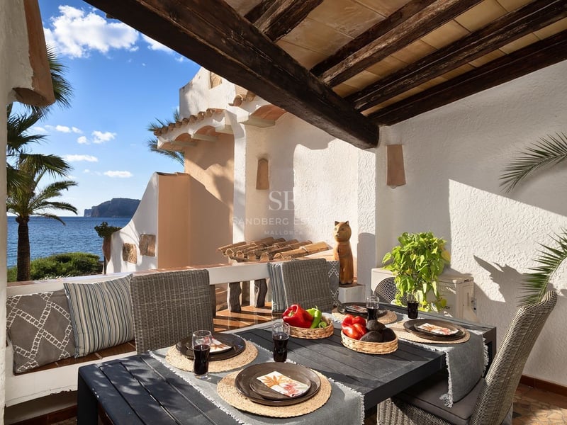 Terrasse ombragée avec table à manger surplombant la mer bleue, palmiers et falaises sous un ciel dégagé.
