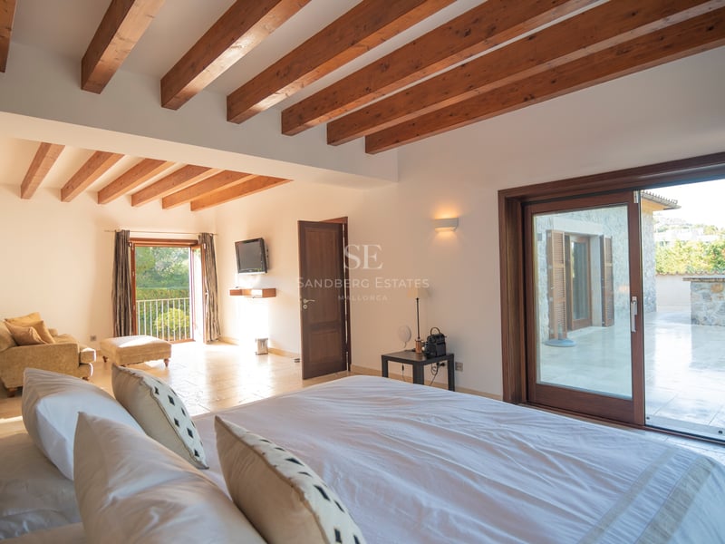 Chambre spacieuse avec hauts plafonds, poutres en bois, sol en pierre et baies vitrées coulissantes menant à la terrasse.