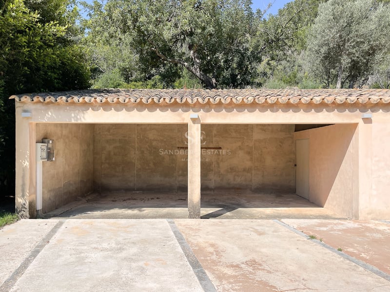 Abri de voiture ouvert à deux places avec toit en tuiles de terre cuite et murs beiges, entouré d'arbres.