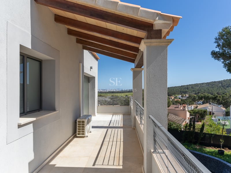 Long balcon couvert avec poutres en bois, murs blancs et garde-corps métallique surplombant une vallée verdoyante.