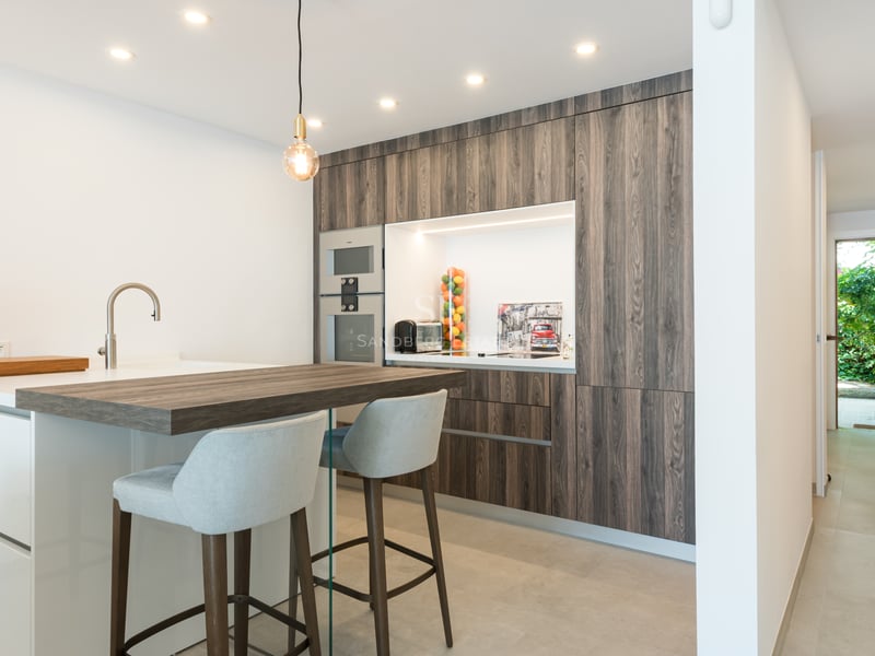 Cuisine contemporaine avec placards en bois sombre, îlot blanc et vue sur l'entrée du jardin au fond du couloir.