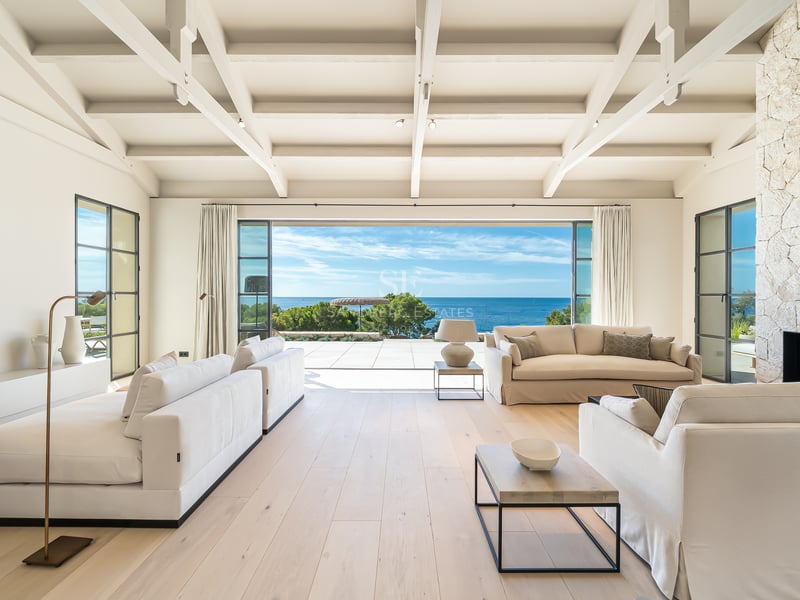 Salon spacieux avec plafonds à poutres blanches, parquet clair et grandes baies vitrées s'ouvrant sur la mer.