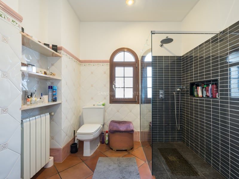 Salle de bain avec sol en terre cuite, carrelage décoratif, fenêtre cintrée en bois et douche moderne vitrée.