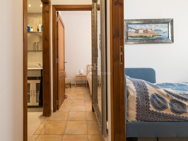 Un couloir avec carrelage en terre cuite menant à une salle de bain et deux chambres, doté d'un grand miroir argenté.
