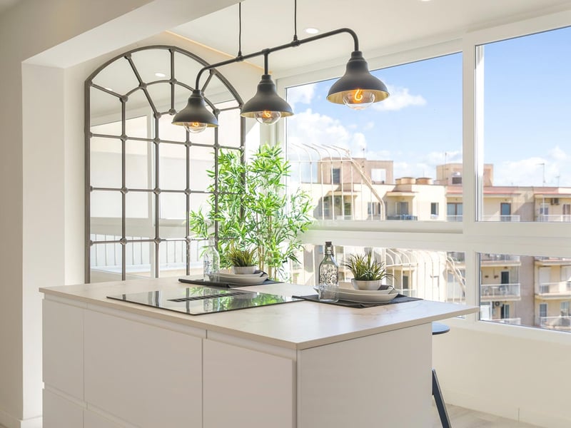 Îlot de cuisine blanc avec plaque à induction, suspensions industrielles et grandes fenêtres vue ville.