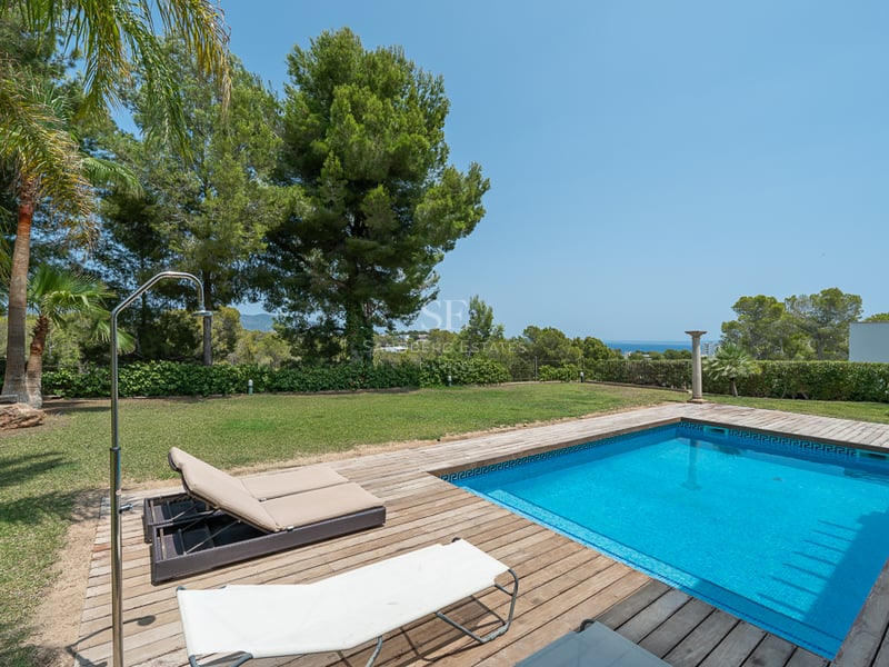 Piscine rectangulaire en mosaïque bleue entourée d'une terrasse en bois et d'une pelouse avec vue mer lointaine.
