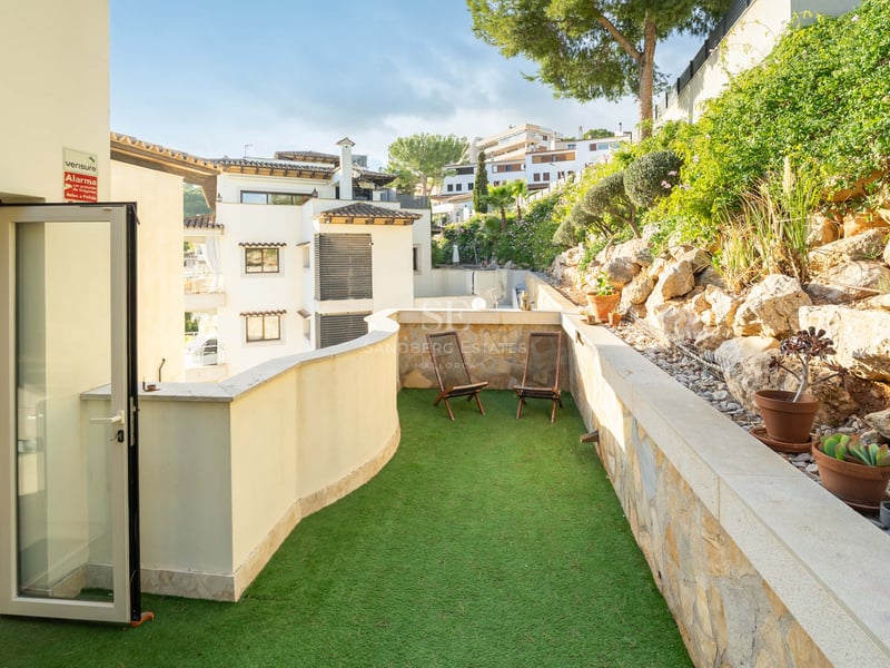 Terrasse ensoleillée avec gazon artificiel vert, deux chaises en bois et un mur en pierre naturelle.