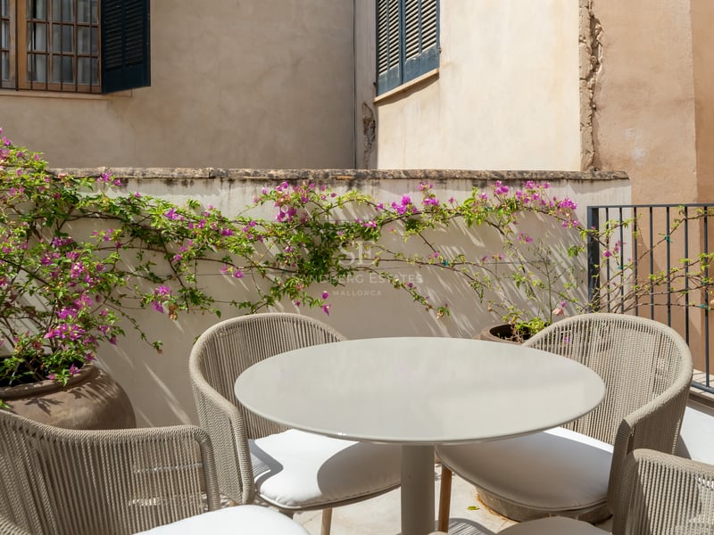 Mesa redonda gris y sillas de cuerda trenzada en una terraza soleada con buganvillas rosas y muros de piedra.