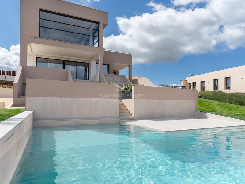 Piscine turquoise au premier plan devant une villa contemporaine beige sous un ciel bleu parsemé de nuages.