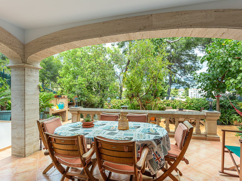 Terrasse en pierre couverte avec table ronde, chaises en bois et balustrade donnant sur un jardin verdoyant.