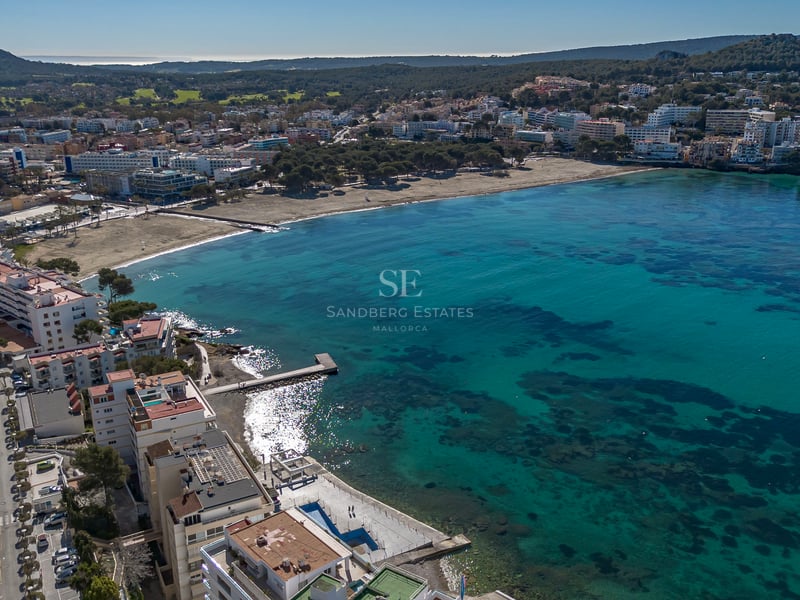 Vue aérienne par drone d'une baie turquoise avec plage de sable et bâtiments côtiers sous un ciel bleu clair.