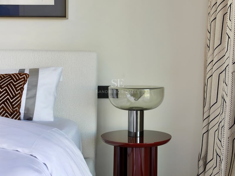 Détail d'une chambre avec tête de lit blanche, table de chevet laquée rouge, lampe en verre et art abstrait.