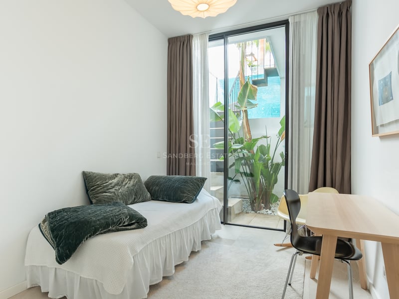 Chambre lumineuse avec un lit simple, un bureau en bois et une porte vitrée ouvrant sur un petit jardin extérieur.