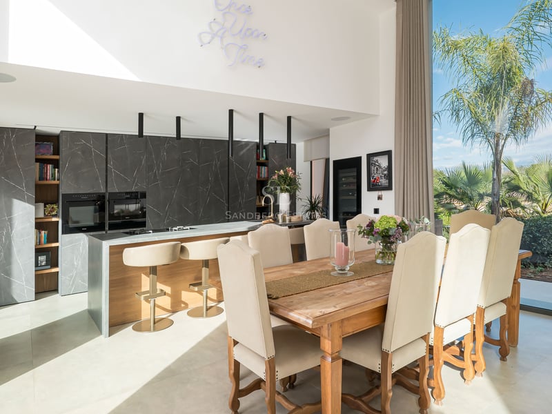 Salle à manger moderne avec table en bois, chaises rembourrées et cuisine en marbre sombre en arrière-plan.