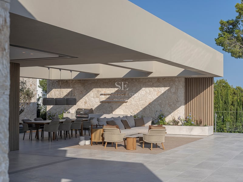 Terrasse de luxe avec salon gris, murs en pierre et espace repas sous un toit architectural moderne.