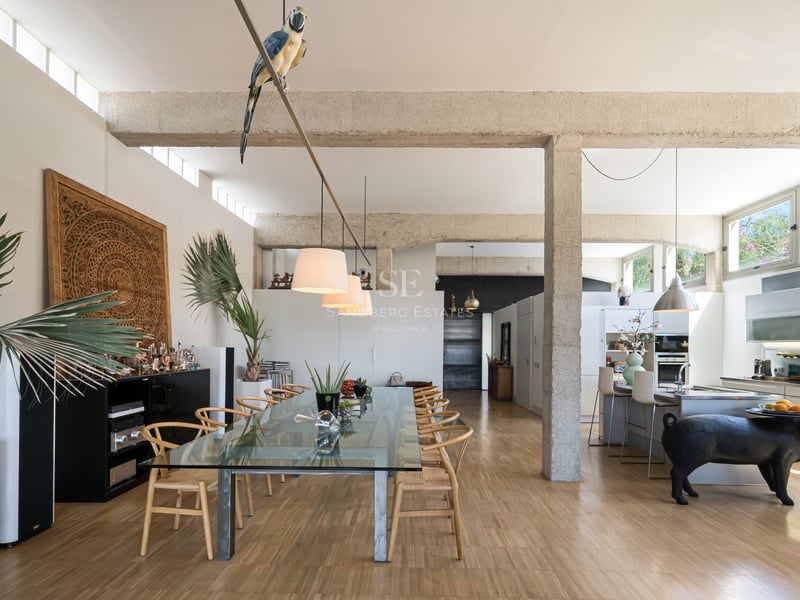 Vaste salle à manger avec poutres en béton brut, table en verre et cuisine intégrée baignée de lumière.