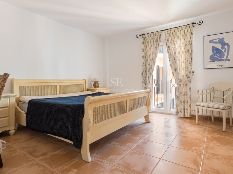 Chambre avec lit en osier, sol en terre cuite, bureau et balcon avec rideaux à motifs floraux.