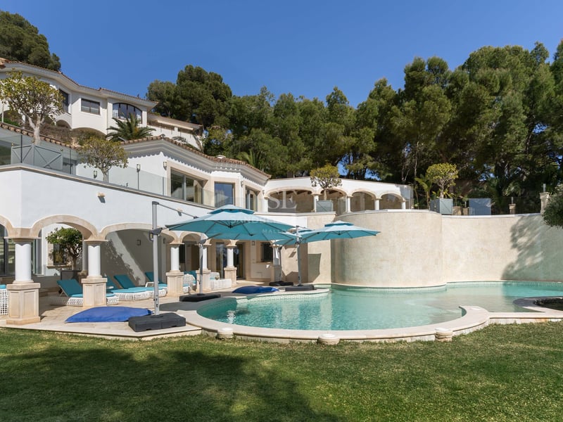 Piscine incurvée avec parasols turquoise à côté d'une villa blanche méditerranéenne entourée de pins.