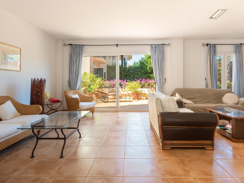 Grand salon avec canapés en osier, sol en terre cuite et grandes baies vitrées ouvrant sur une terrasse ensoleillée.