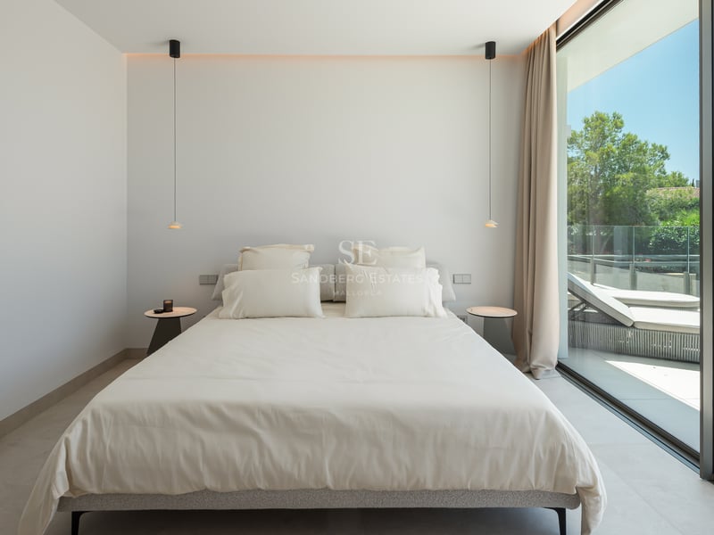 Chambre minimaliste avec grand lit, suspensions design et baies vitrées ouvrant sur une terrasse ensoleillée.
