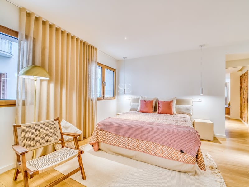 Chambre lumineuse avec lit double, textiles roses, parquet en bois, fauteuil en osier et fenêtres avec rideaux.