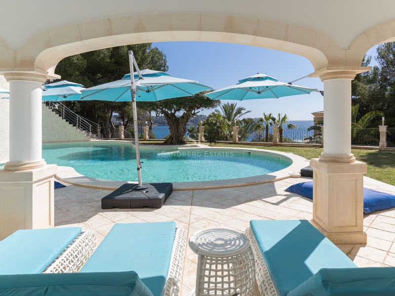 Une piscine turquoise incurvée entourée de dalles en pierre et de chaises longues turquoises sous une arche ombragée.