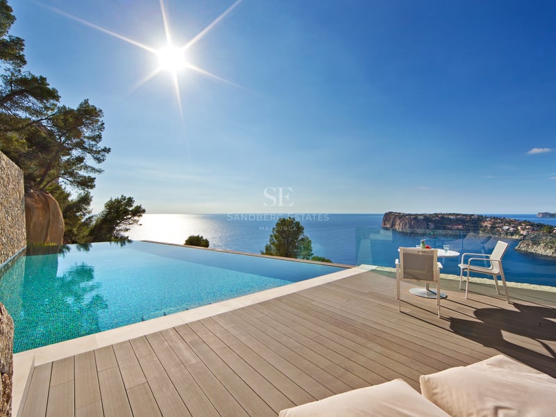 Piscine à débordement turquoise sur terrasse en bois avec vue sur mer bleue et côte rocheuse sous le soleil.