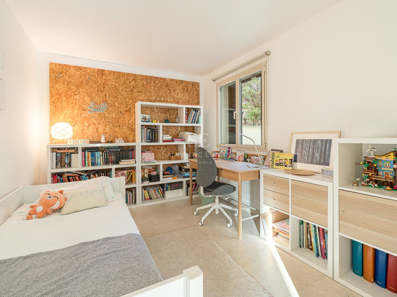 Chambre d'enfant avec lit blanc, bureau en bois et étagères remplies de livres sous une fenêtre ensoleillée.