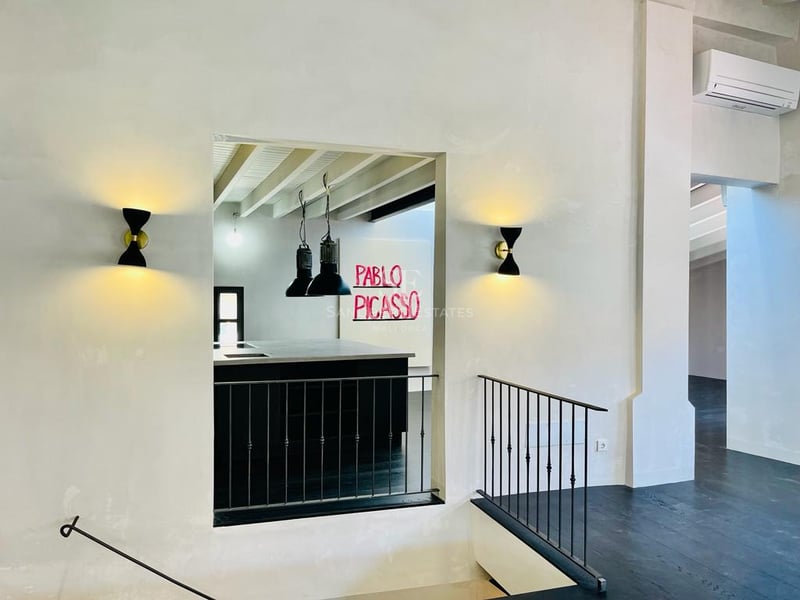 Vue à travers une ouverture murale montrant un îlot de cuisine, des suspensions et une rampe en métal noir.