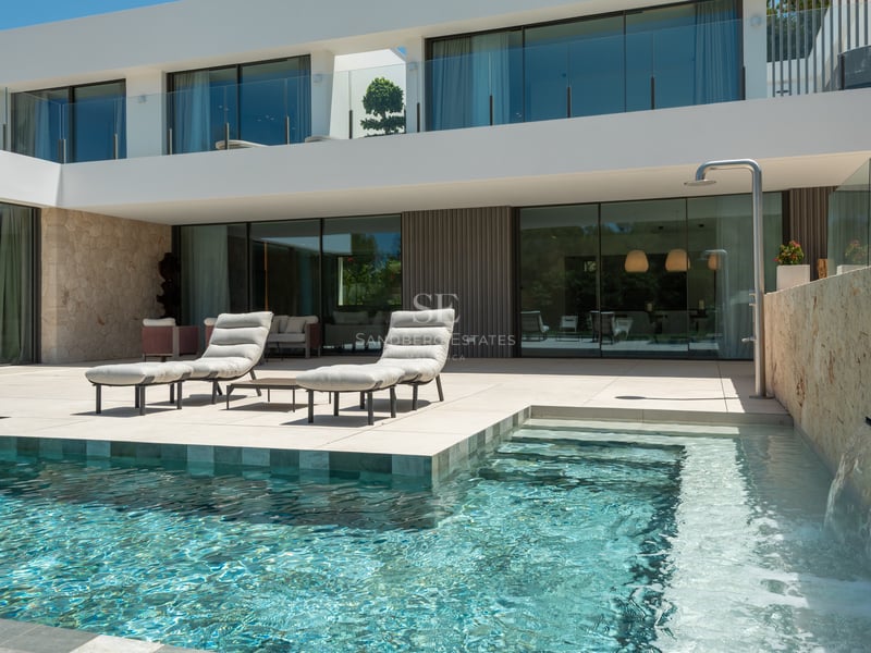 Terrasse de villa moderne avec piscine turquoise, cascade en pierre, bains de soleil et baies vitrées.
