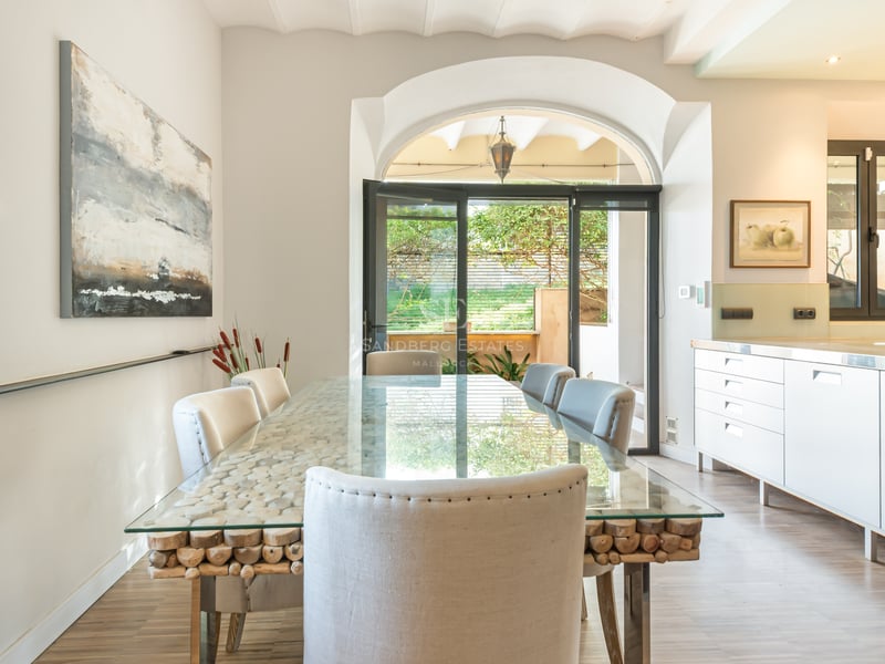 Salle à manger lumineuse avec table en verre sur socle en bois, chaises blanches et arche menant à l'extérieur.