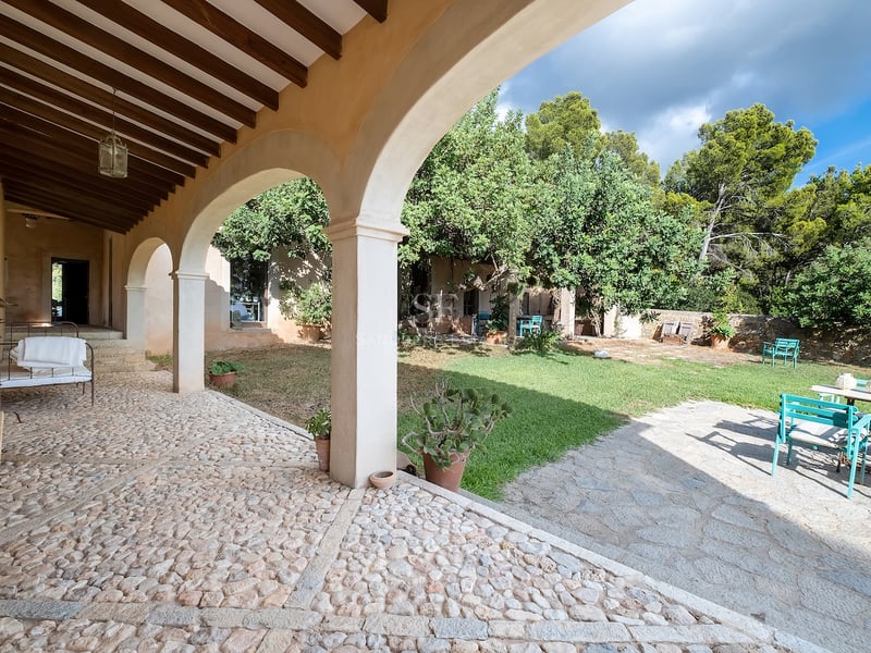 Porche traditionnel aux arches de pierre avec sol en pavés donnant sur un jardin ensoleillé avec meubles turquoise.