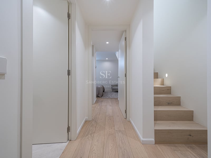 Couloir lumineux avec parquet en chêne clair, murs blancs et escalier en bois avec éclairage encastré.