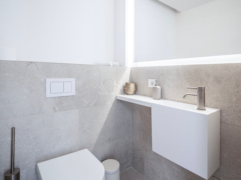 Salle de bain minimaliste avec WC blanc, petit lavabo suspendu et grand miroir rétroéclairé sur murs en pierre grise.