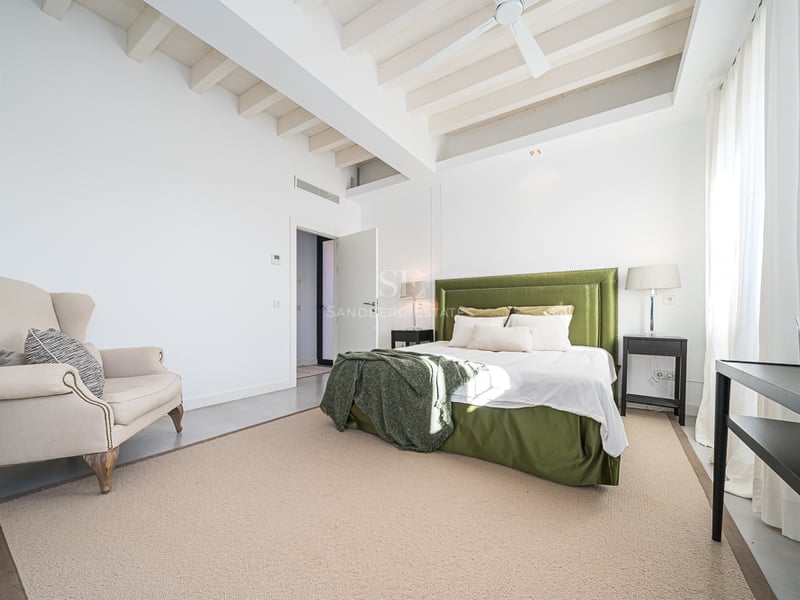 Chambre lumineuse avec poutres blanches, lit en velours vert, fauteuil beige et grandes fenêtres.