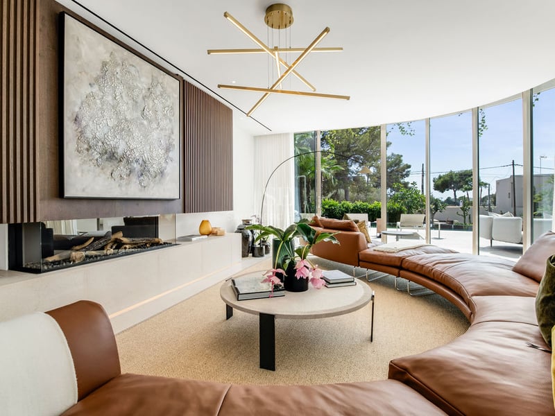 Salon moderne avec canapé courbé en cuir cognac, cheminée linéaire et grandes baies vitrées donnant sur un jardin.