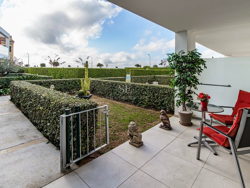 Terrasse blanche moderne avec deux chaises rouges et une table donnant sur un jardin privé aux haies taillées.