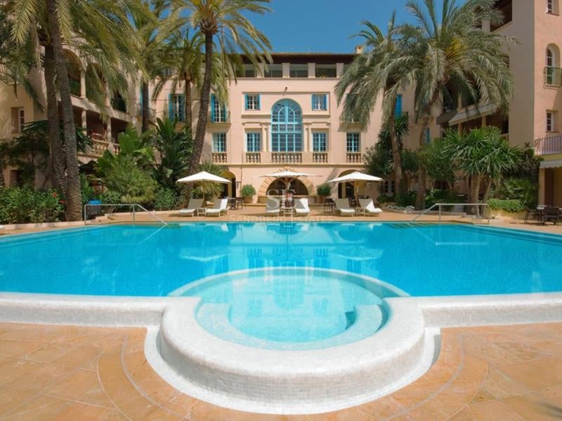 Grande piscine turquoise avec entrée circulaire, entourée de palmiers et d'un bâtiment classique de couleur pêche.