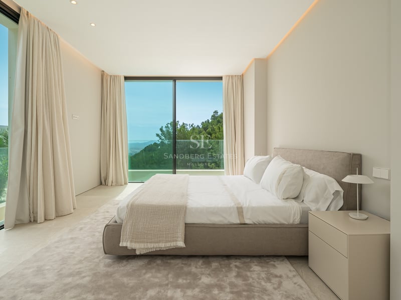 Chambre spacieuse avec lit double, grandes baies vitrées donnant sur un paysage verdoyant et des rideaux clairs.