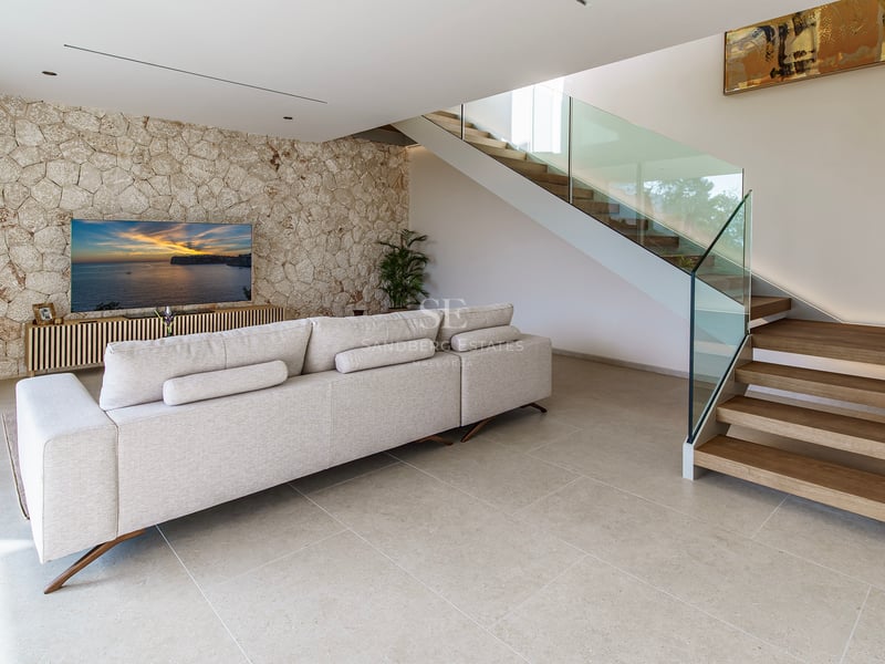Salon spacieux avec canapé beige, mur en pierre décoratif, télévision et escalier en bois avec garde-corps en verre.