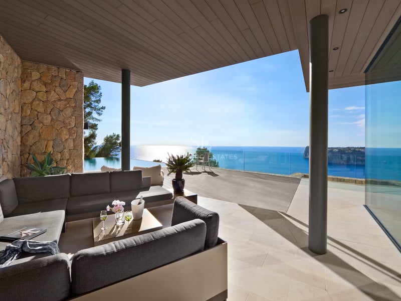 Salon extérieur avec canapés gris sur une terrasse en pierre surplombant une piscine à débordement et la mer.