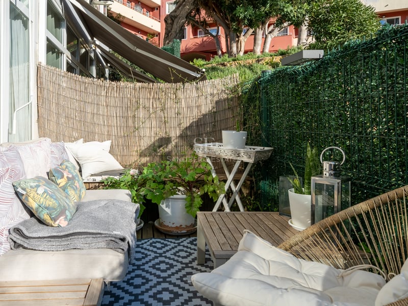 Terrasse ensoleillée avec canapé confortable, tapis géométrique et brise-vue en bambou dans un cadre côtier.