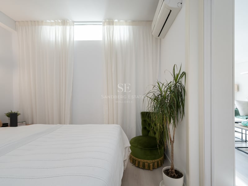 Chambre blanche épurée avec lit, fauteuil en velours vert, plante verte et vue sur le salon attenant.