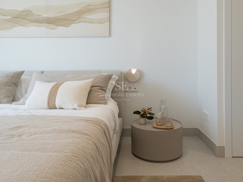 Cama con ropa de lino beige junto a una mesita de noche minimalista circular y arte abstracto en la pared.