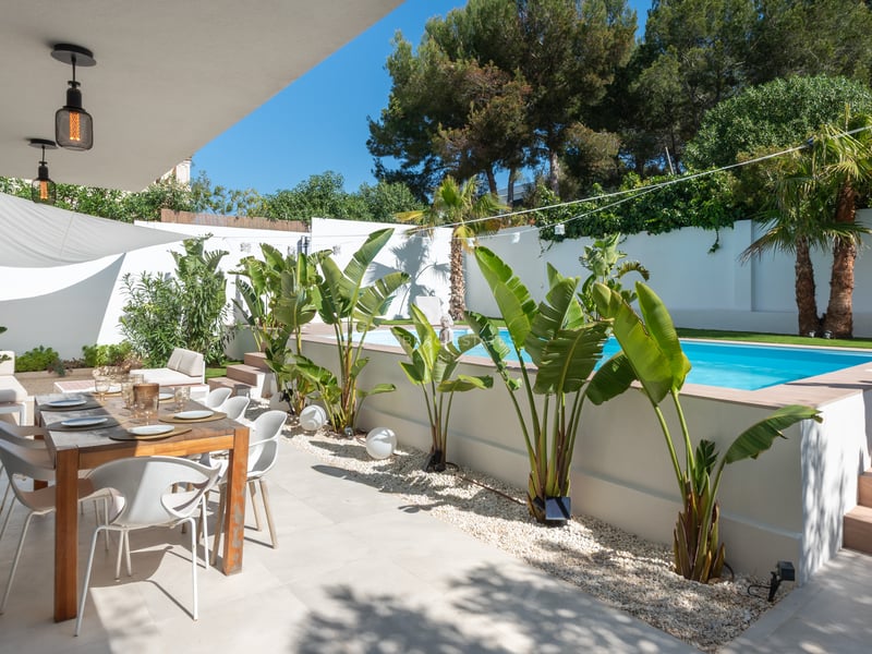 Une terrasse moderne avec table à manger en bois sous un auvent à côté d'une piscine surélevée et de plantes tropicales.