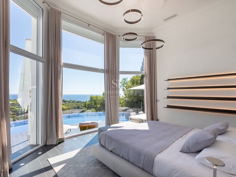 Chambre moderne avec de grandes baies vitrées donnant sur une piscine à débordement et la mer Méditerranée.
