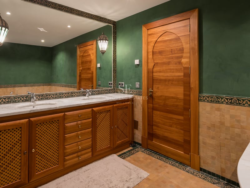 Salle de bain aux murs verts, meubles en bois sculpté, double vasque en marbre et grand miroir décoré.
